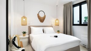 A modern hotel room with a large bed, two bedside lamps, a wall-mounted decorative item, a small plant, and a window with curtains. - Seasalt Luxury Villas (Pefkochori)