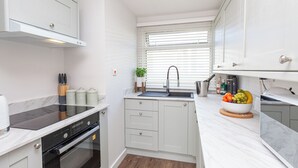 A compact kitchen with white cabinetry, a black induction cooktop, a built-in oven, and a window with blinds.