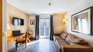 Une chambre d’hôtel avec un bureau en bois, un ordinateur portable, un canapé en cuir marron, un téléviseur fixé au mur, un tableau encadré et une porte-fenêtre coulissante donnant sur le balcon.