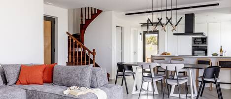 Uma sala de estar moderna com um sofá seccional cinza, uma escada de madeira e uma área de cozinha com mesa e cadeiras.