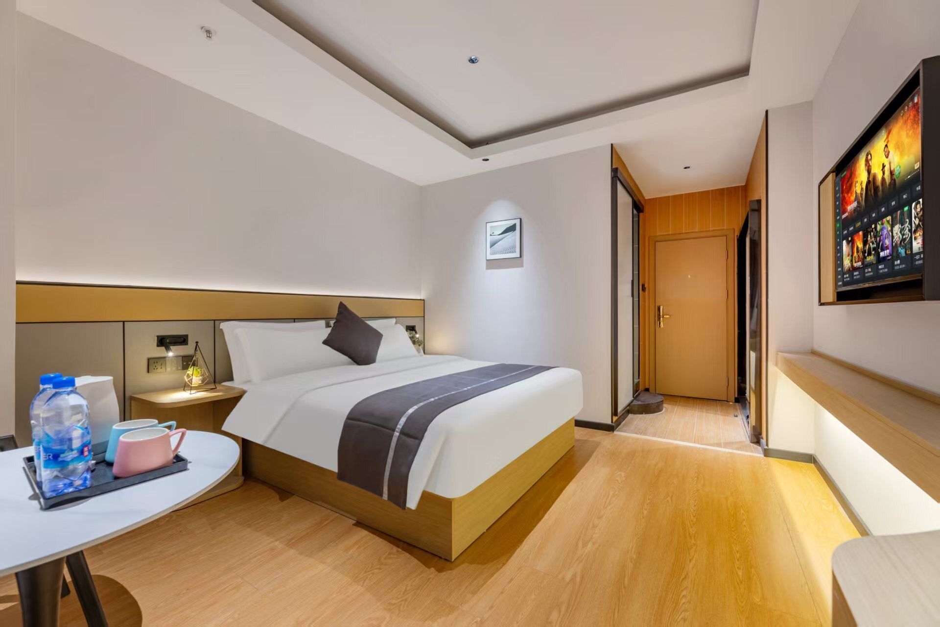 Quarto de hotel moderno com uma cama grande, mesa de cabeceira com abajur, pequena mesa com garrafas de água e chávenas, e uma TV fixada na parede.