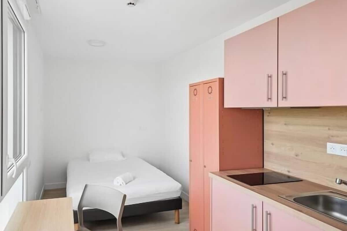 Een kleine kamer met een bed, een tafeltje, een stoel en een kitchenette met roze kastjes, plus een raam.
