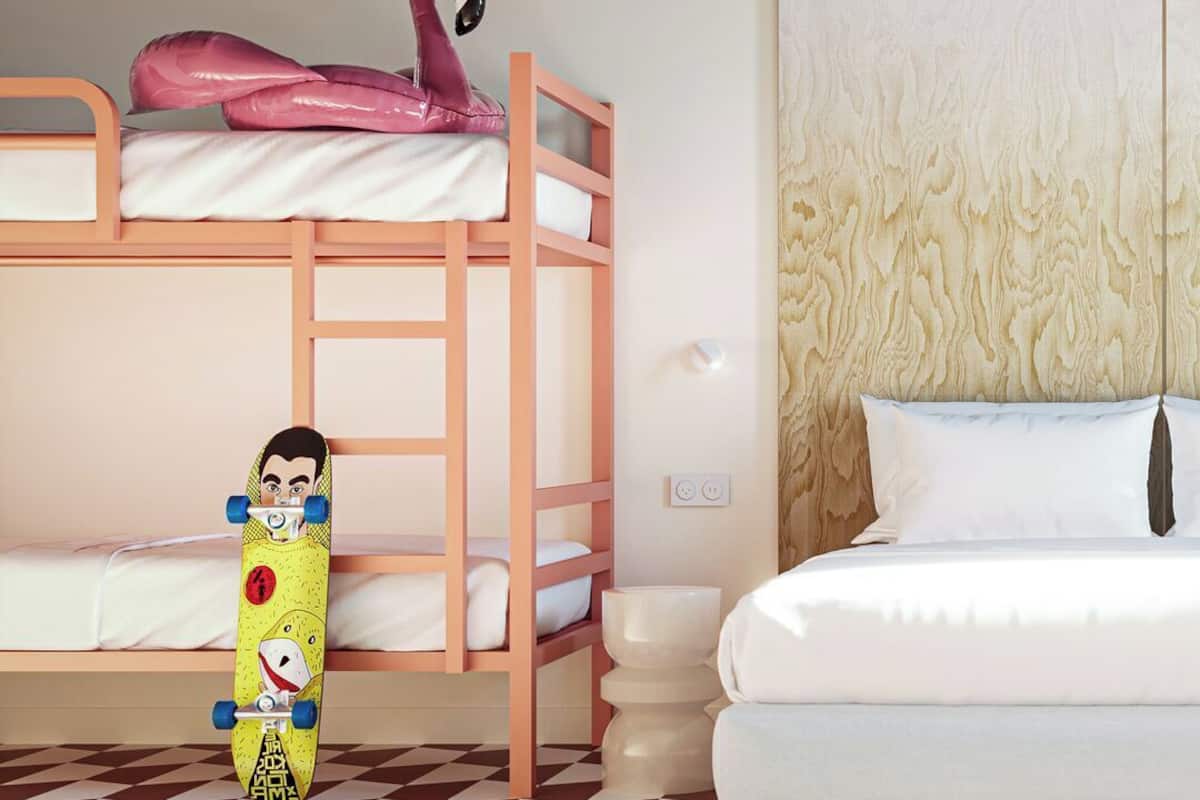 Een stapelbed met een roze flamingo decoratie, een geel skateboard met tekenfilmfiguren, en een witte bank met een behangetje op de muur.