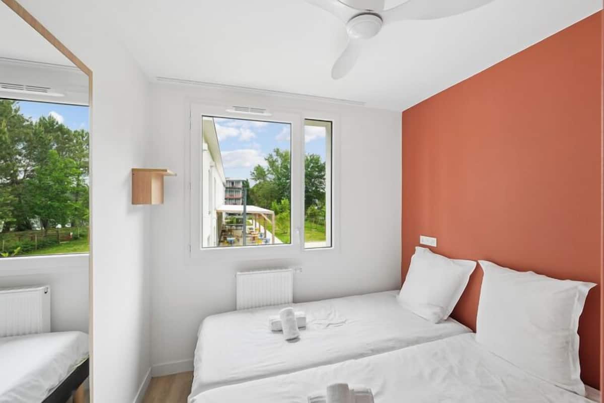 Een slaapkamer met twee bedden, een raam, een plafondventilator en uitzicht op groen.