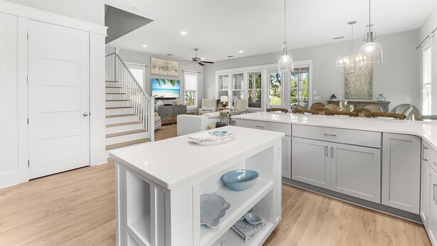 Une cuisine moderne avec des meubles blancs, un îlot central, des luminaires suspendus et une vue sur le salon, avec un escalier.