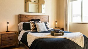 A neatly made bed with a wooden headboard, two bedside tables with lamps, a mirror above the bed, and a window with curtains. - No Fee | New Private Cottage | Fireplace + Full Kitchen (Post Falls)