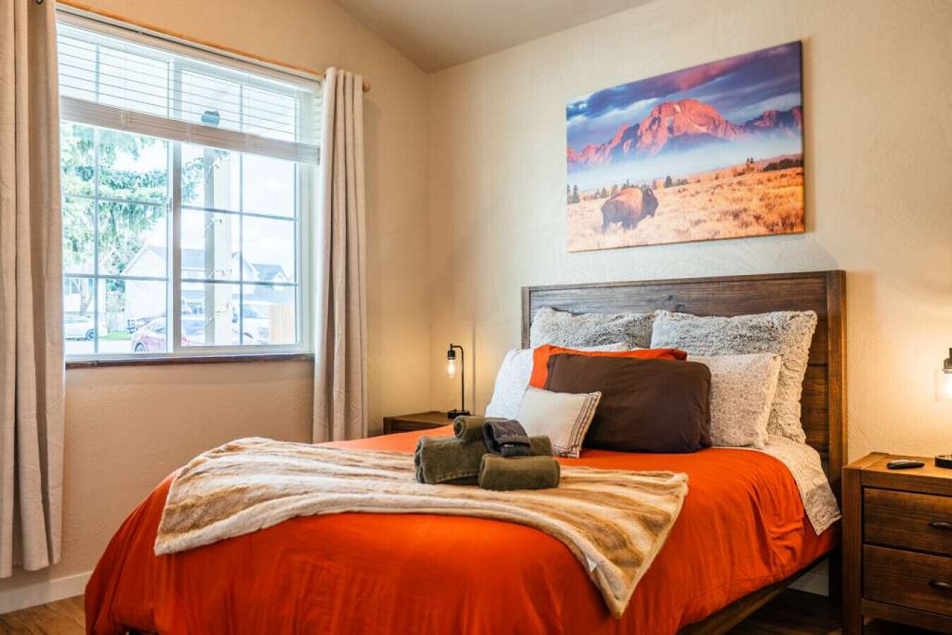 Une chambre à coucher avec un lit, une table de chevet, une lampe, un tableau et une fenêtre avec des rideaux.