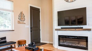 A living room with a fireplace, a flat-screen TV mounted above it, a coffee table with a remote control, and a door leading to another room. - No Fee | New Private Cottage | Fireplace + Full Kitchen (Post Falls)
