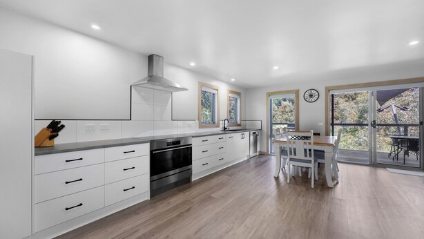A modern kitchen with white cabinetry, stainless steel appliances, and a dining area with a table and chairs. - Alpine Edge Retreat - Gateway to Mount Buller (Merrijig)