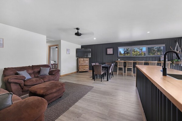 A modern living room with a brown sofa, a wooden dining table with chairs, and a kitchen area with a black countertop.