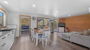A modern kitchen with white cabinets and a wooden countertop, a dining area with a wooden table and white chairs, a grey sofa, and a TV on a wooden wall. - Alpine Edge Retreat - Gateway to Mount Buller (Merrijig)