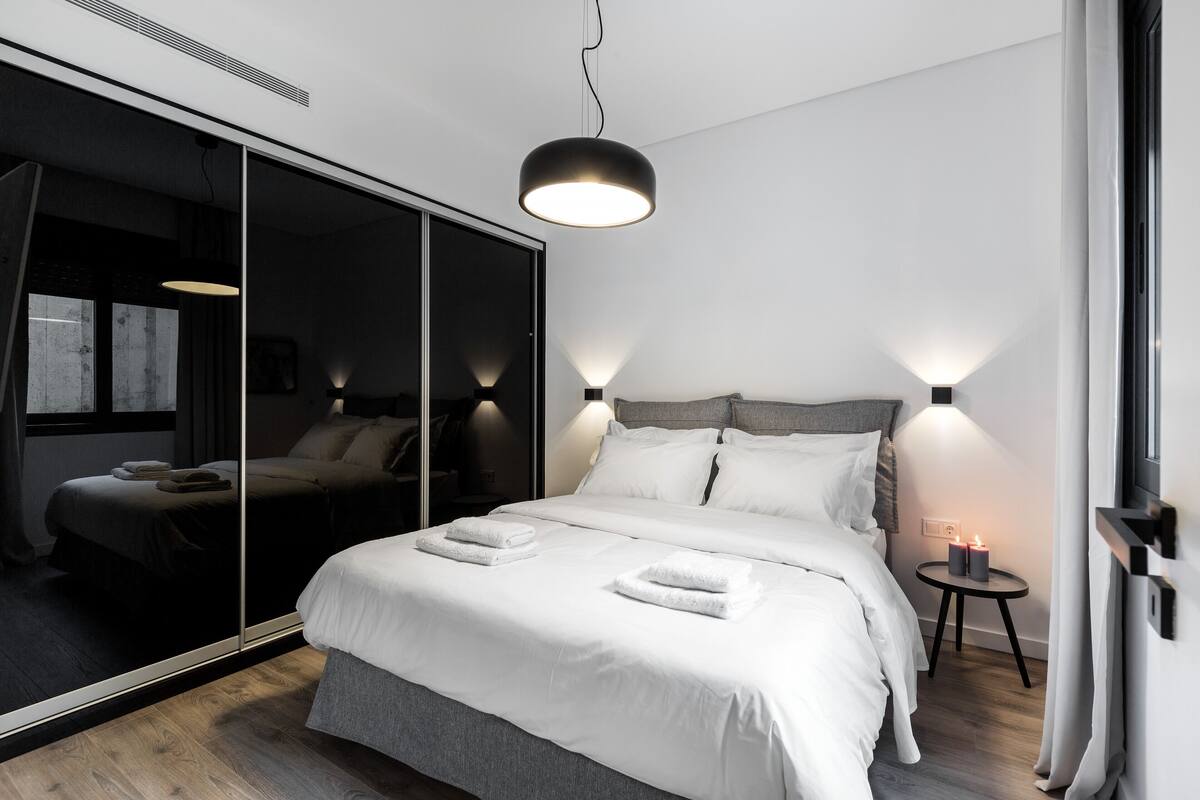 Un dormitorio moderno con un espejo grande, una cama con ropa de cama blanca, dos mesitas de noche con velas y una lámpara colgante.