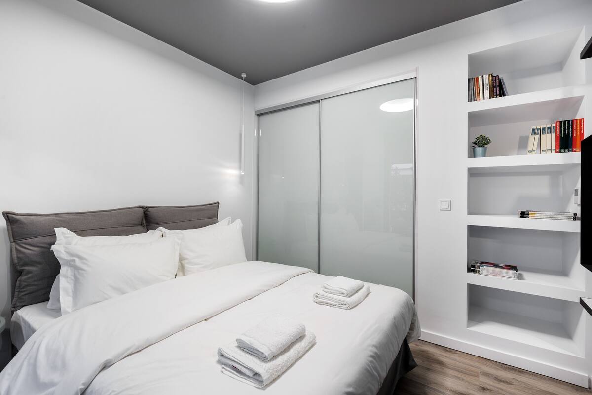 Una habitación de hotel moderna con una cama, ropa de cama blanca, un cabecero gris, una estantería empotrada y un armario con puerta corredera.