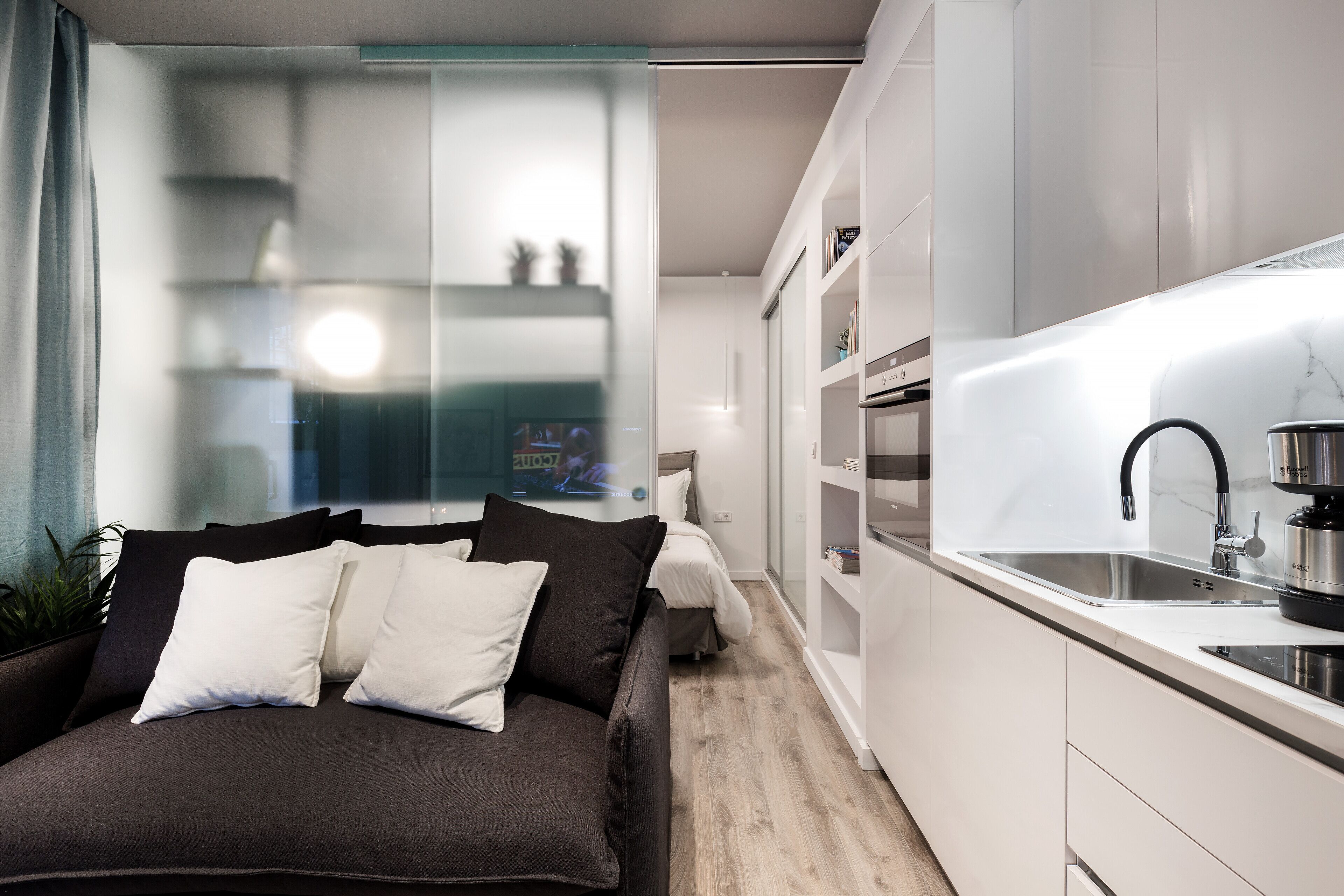 A modern, compact living space with a sofa, a kitchen area with a sink and stove, and a glass partition separating the living area from a bedroom.