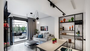 A modern living room with a sofa, coffee table, and dining area. There is a bookshelf with decorative items and a large window offering a view of greenery. - Elysan Urban Stays (Athens)