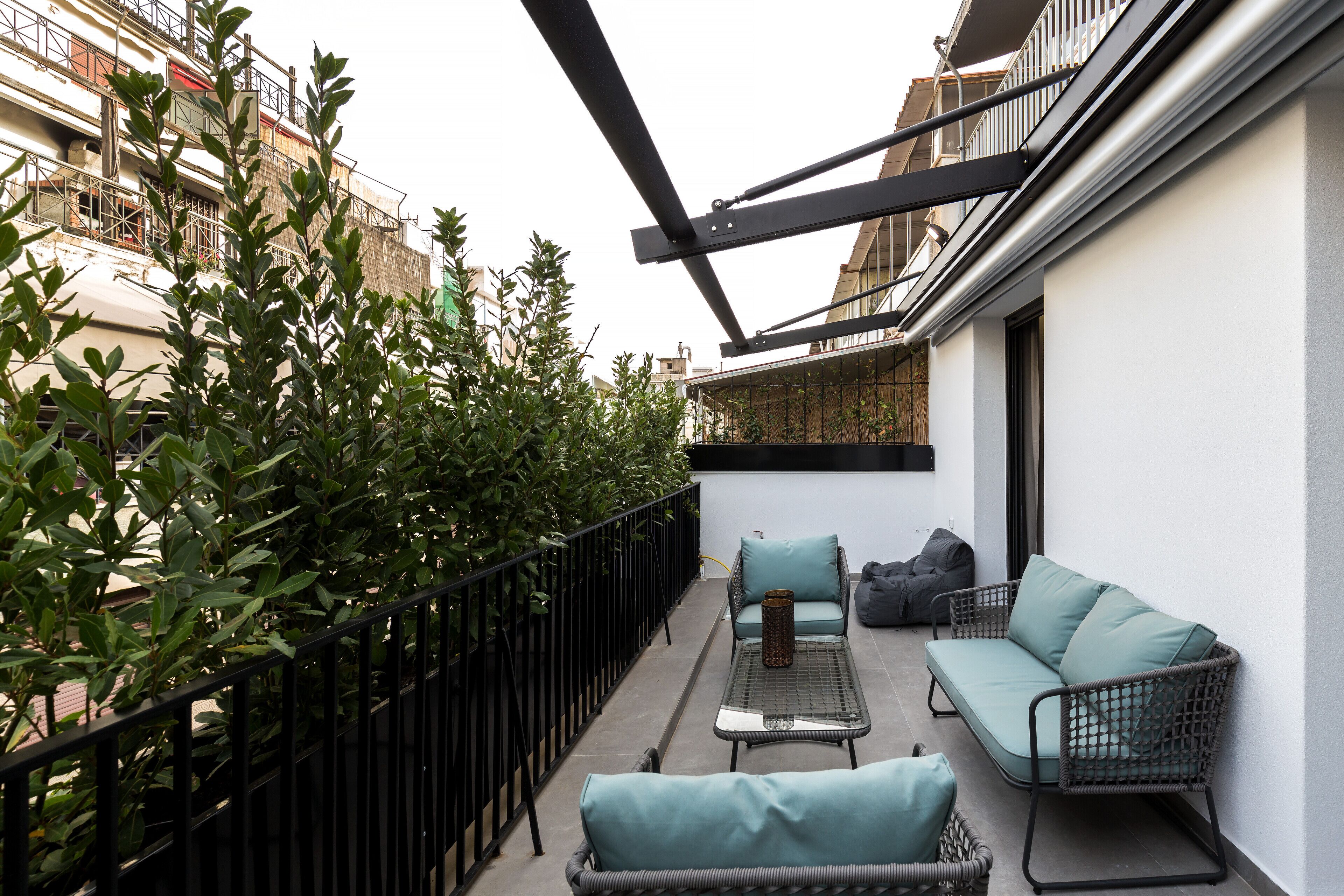 Ein Balkon mit einer Couch, einem Tisch und einem Stuhl. Ein Geländer und etwas Grün sind ebenfalls vorhanden.