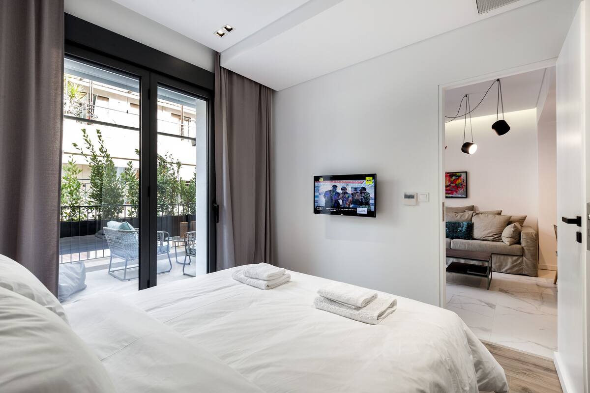 Una habitación de hotel moderna con una cama grande, un televisor montado en la pared y un balcón con mobiliario exterior.