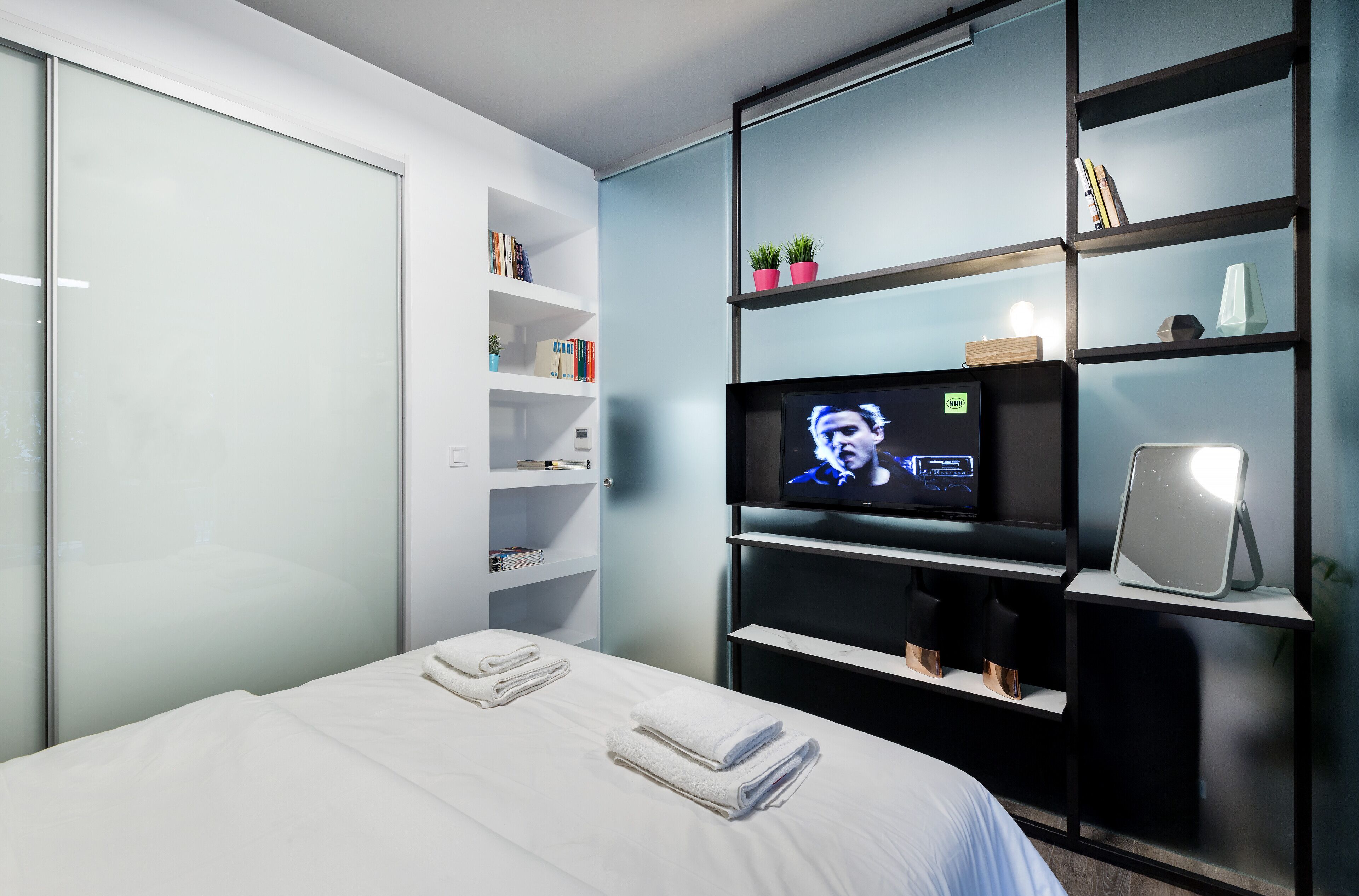 A modern hotel room with a bed, white linens, a television, a bookshelf, and a desk with a lamp.