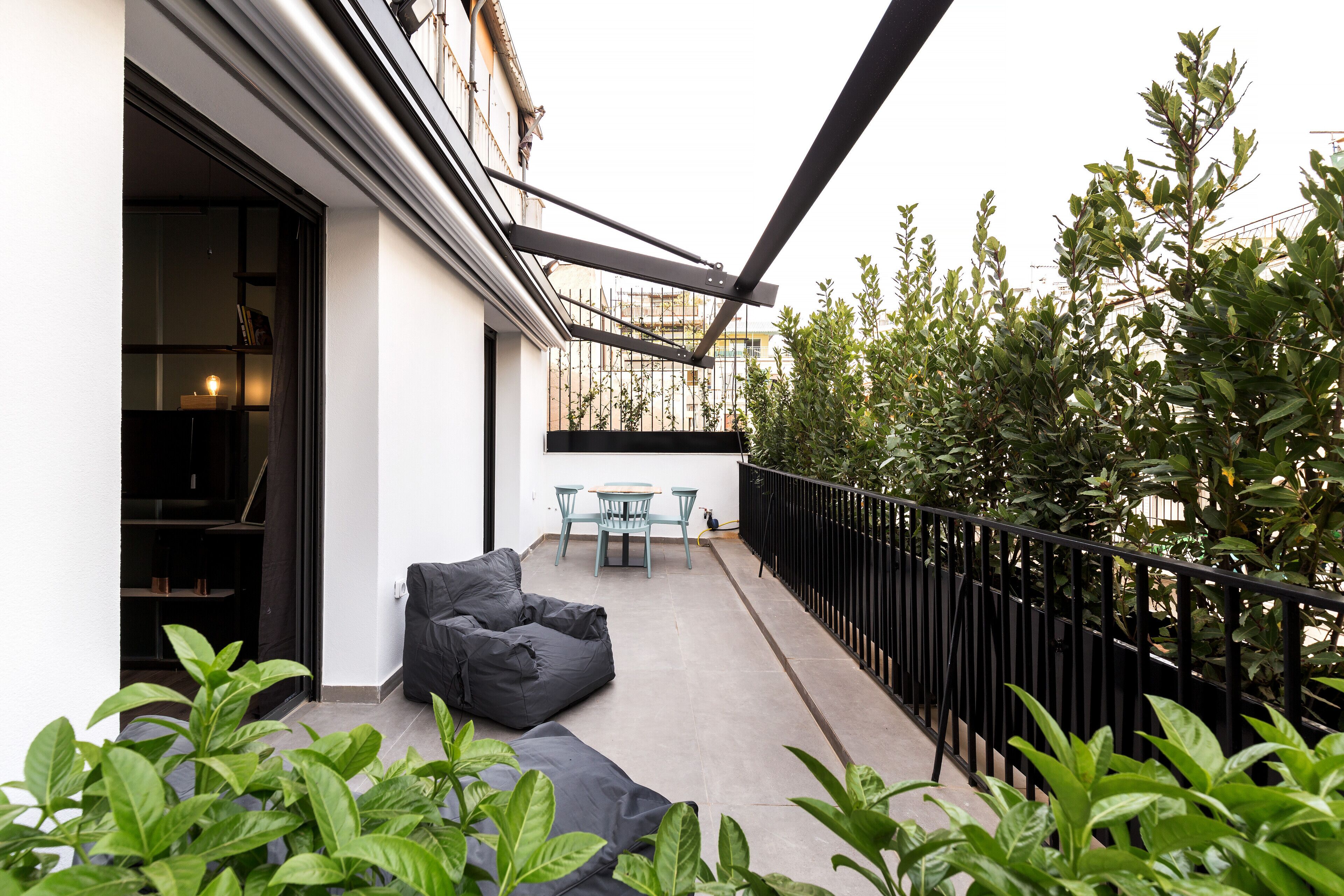 A balcony with a black metal railing, a black bean bag chair, a small table with two chairs, and a view of a building with a window and a door.