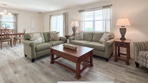 Ein Wohnzimmer mit zwei Sofas, einem Couchtisch und zwei Lampen. Im Hintergrund steht ein Esstisch mit Stühlen.