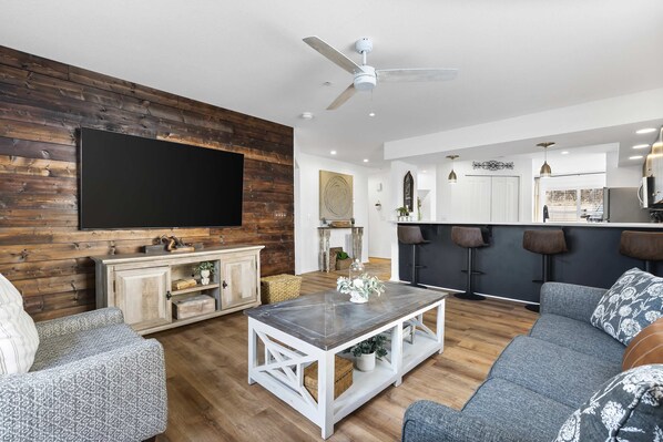 A modern living room with a flat-screen TV, a ceiling fan, and a bar area with stools.
