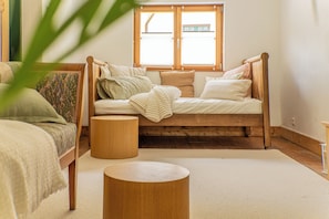 Un dormitorio con una cama de madera, dos mesitas de noche redondas de madera, una ventana con persianas y una planta grande al frente.