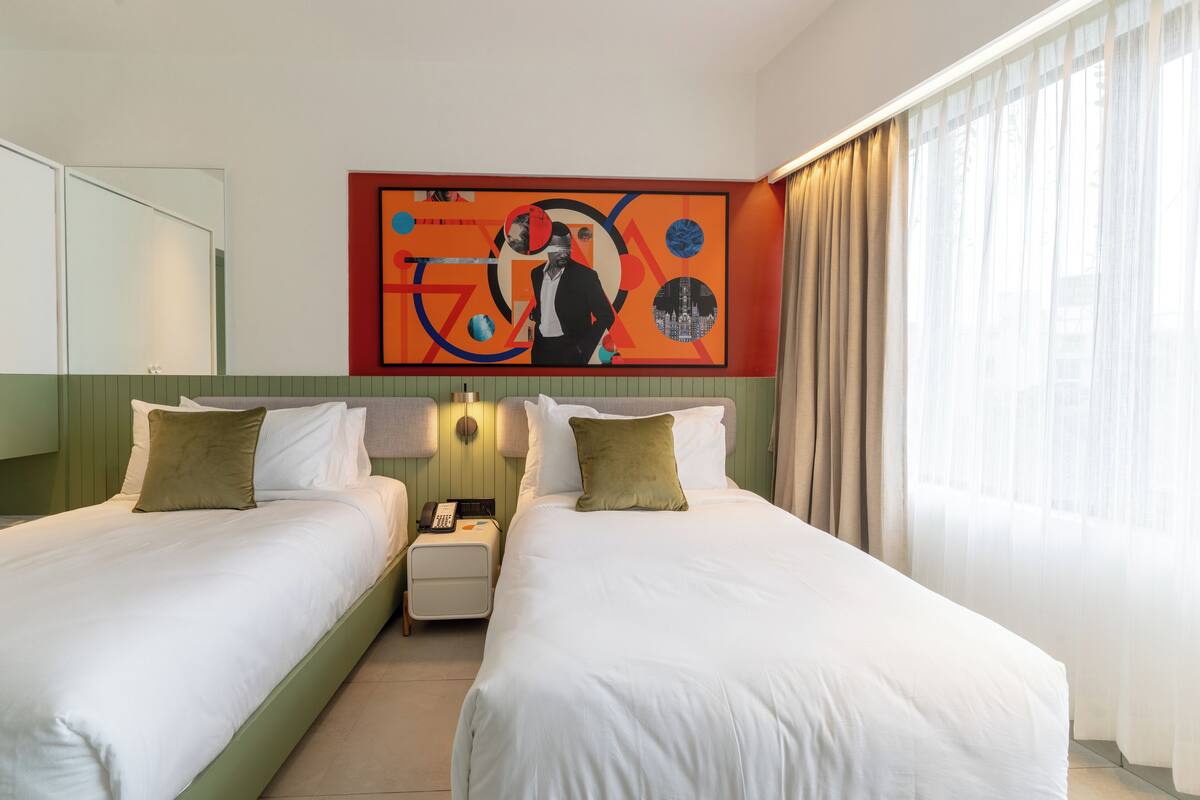 Ein Hotelzimmer mit zwei Betten, einem Nachttisch mit Telefon, einem Wandbild und einem Fenster mit Vorhängen.
