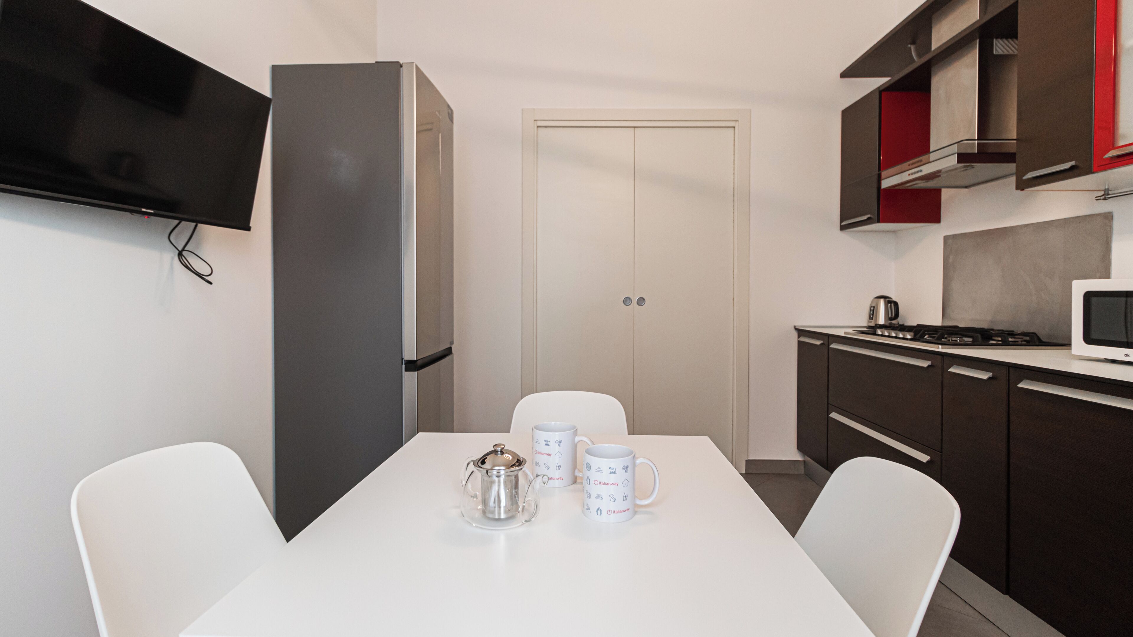 A compact kitchen with a refrigerator, microwave, and stove. There is a dining table with chairs and two coffee mugs.