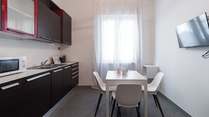 Une cuisine compacte équipée d’un micro-ondes, d’un évier et de placards, une table à manger avec des chaises, et un téléviseur fixé au mur.