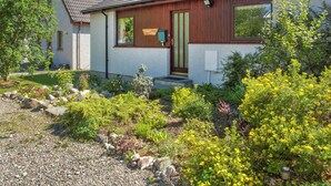 Une maison à deux étages avec une porte d’entrée en bois, un petit porche et un jardin où poussent diverses plantes et fleurs.