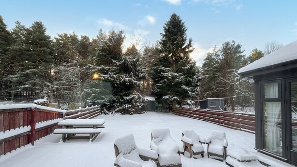 Un espace extérieur aménagé avec des sièges, recouvert de neige, comprenant des chaises en bois et une table de pique-nique, le tout entouré d’arbres chargés de neige et d’une clôture en bois.