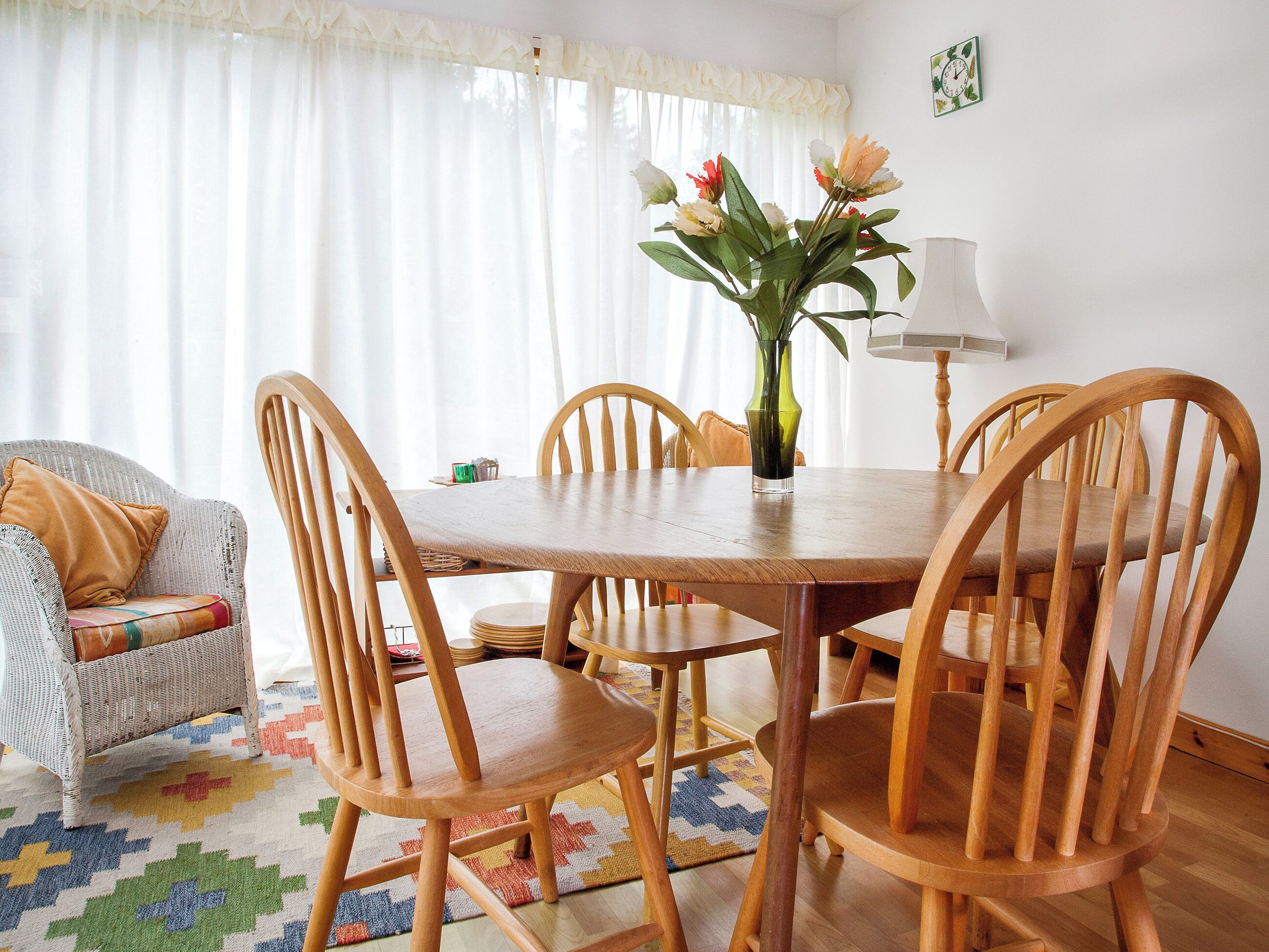 A dining area with a wooden table, six chairs, a vase with flowers, a lamp, and a patterned rug.