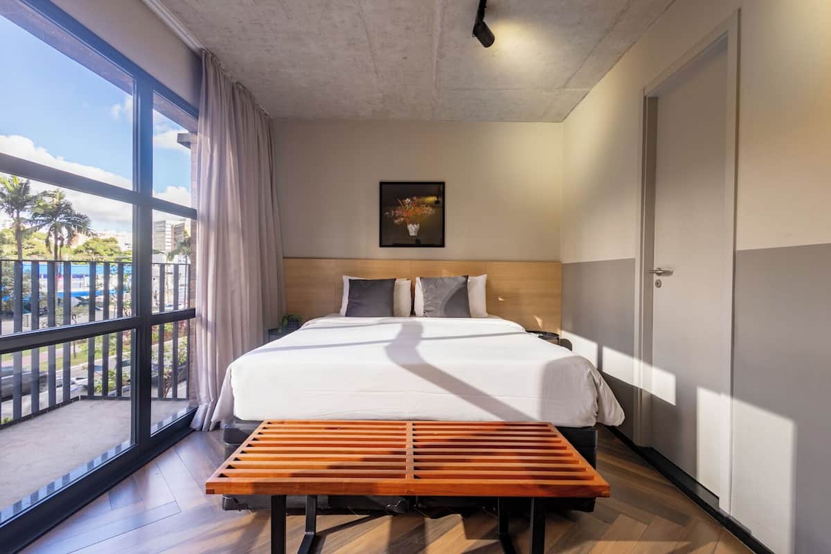 A hotel room with a large bed, a wooden bench, a view of palm trees, and a painting on the wall.