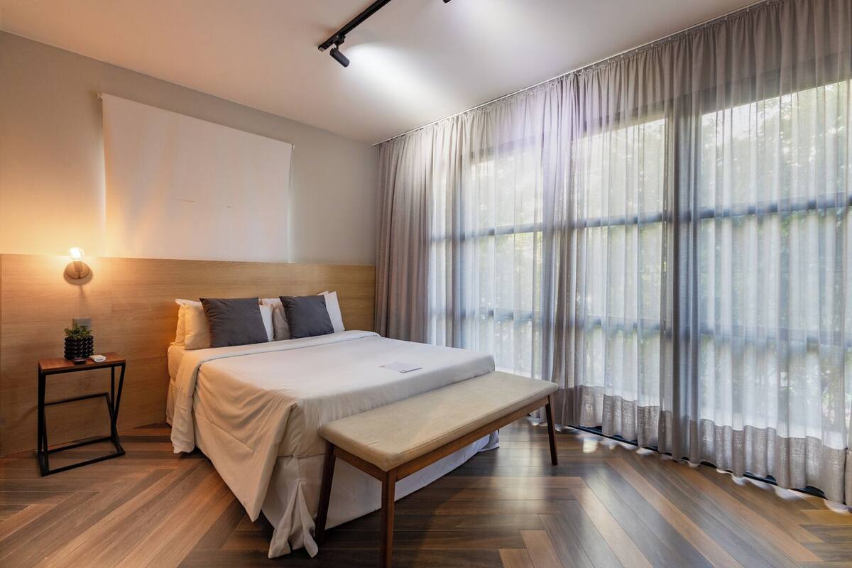 A modern hotel room with a wooden headboard, a large bed with a bench, a bedside table with a lamp, and sheer curtains allowing natural light.