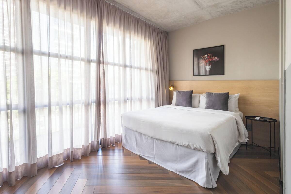 A hotel room with a large bed, wooden flooring, sheer curtains, a headboard, a bedside table, and a framed picture on the wall.