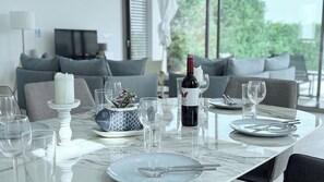 Una mesa de comedor dispuesta con platos, vasos, una vela y una botella de vino, con un sofá y televisor al fondo.