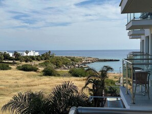 A balcony with a view of the sea, a grassy area, and some buildings in the distance.