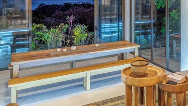 Een ruime kamer met een groot raam, een houten bankje en een ronde houten tafel.