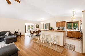 A modern kitchen with wooden cabinets, a bar area, and a dining space with chairs. - Whispering Pines Oasis 12 ac Scenic Forest Retreat (Amherst)