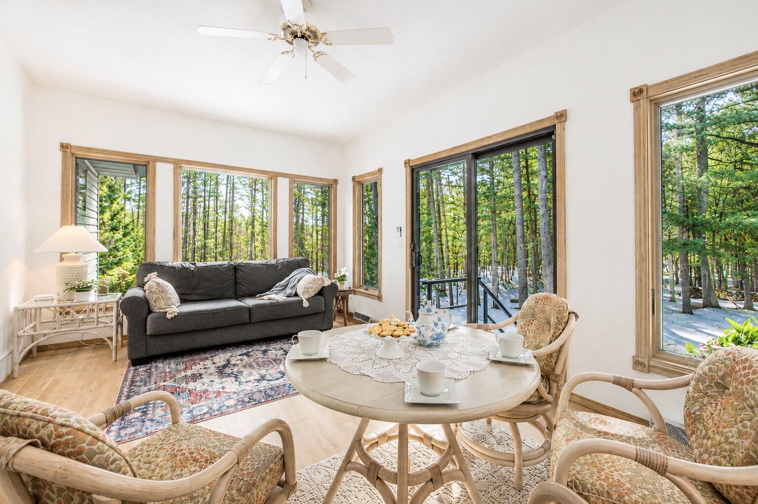 A room with a sofa, a round table with a lace tablecloth, wicker chairs, and large windows with a view of trees.