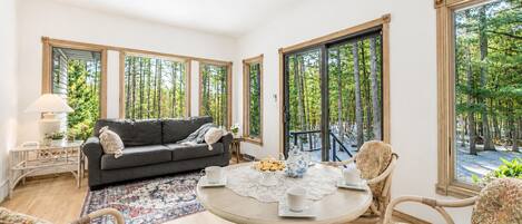 A room with a sofa, a round table with a lace tablecloth, wicker chairs, and large windows with a view of trees.