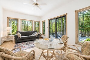 A room with a sofa, a round table with a lace tablecloth, wicker chairs, and large windows with a view of trees. - Whispering Pines Oasis 12 ac Scenic Forest Retreat (Amherst)