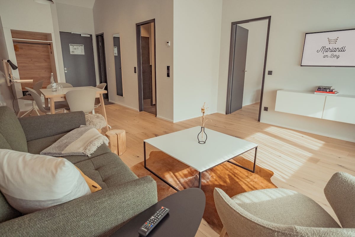 A modern living room with a sofa, a coffee table, and a TV. There is a dining area with a table and chairs. The room has wooden flooring and a large window allowing natural light.