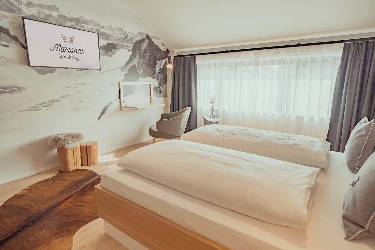 A bedroom with a bed, a chair, a small table, and a wall-mounted TV displaying "Mariandl am Berg".