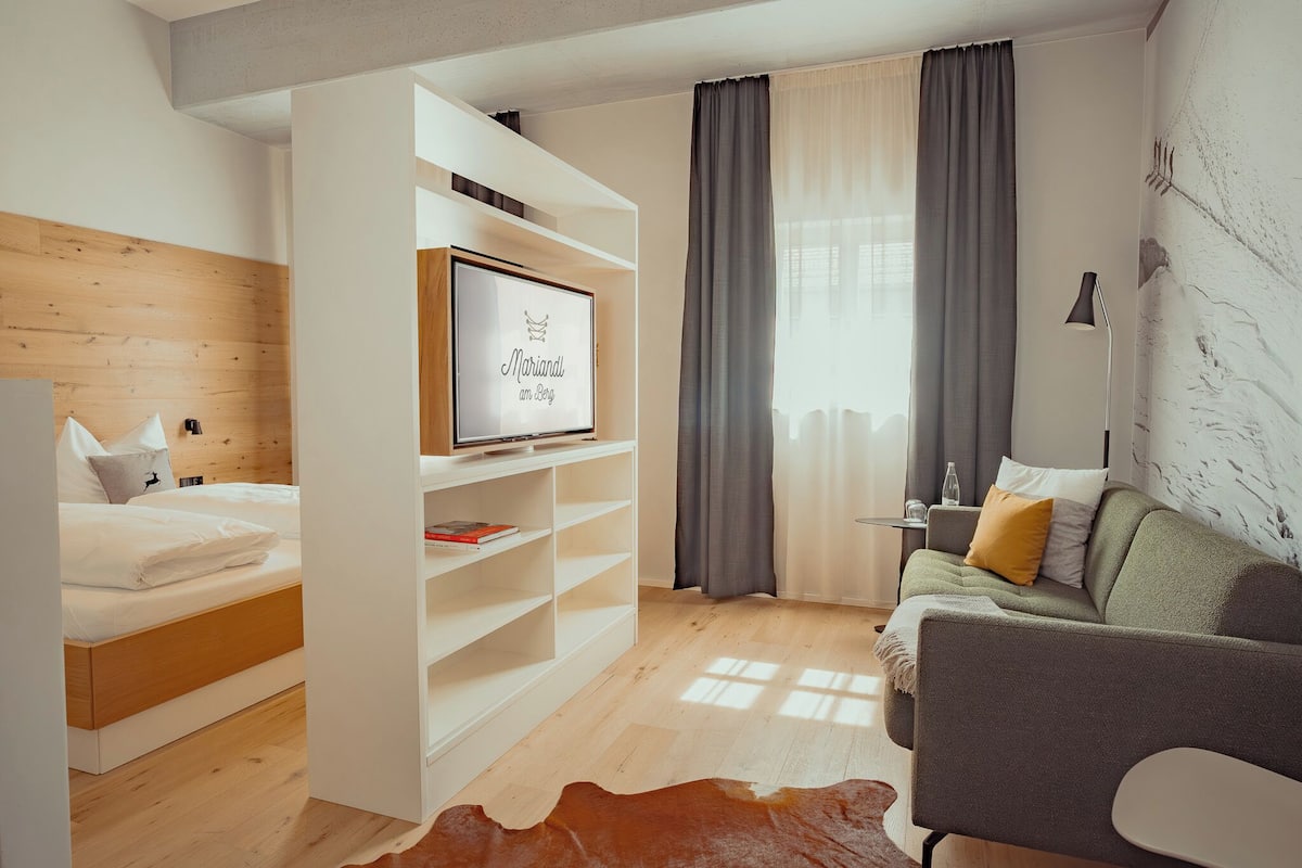 A modern bedroom with a wooden headboard, a bed with white bedding, a white shelving unit with a framed picture, a grey sofa with cushions, and a cowhide rug.