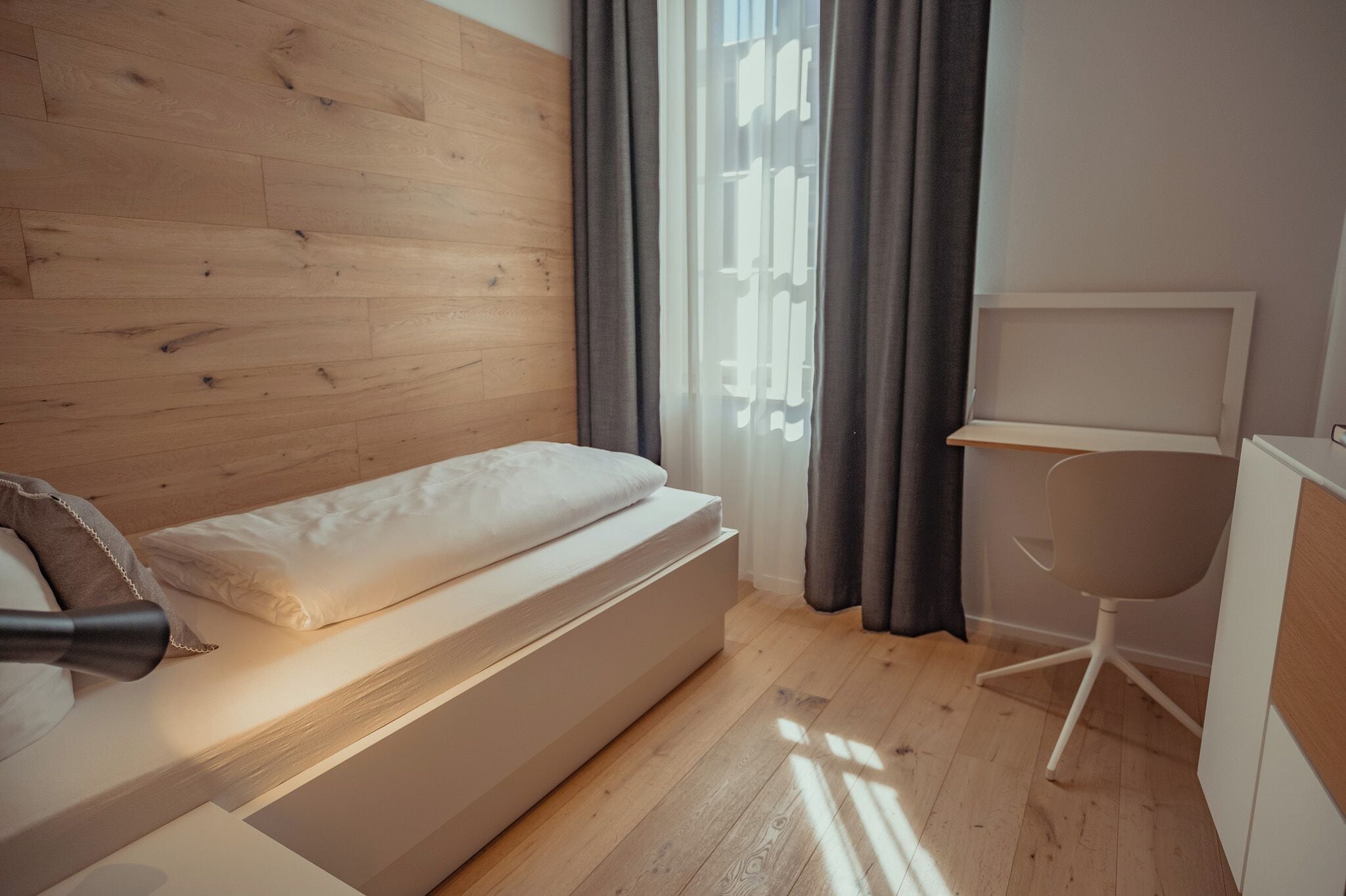 Een moderne slaapkamer met een houten hoofdbord, een bed met matras en kussens, een bureau met stoel, en een raam met gordijnen.