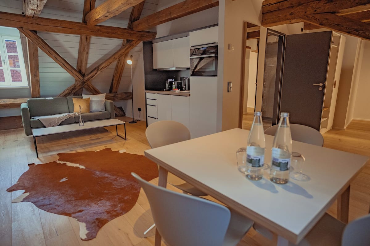 A modern interior with a dining table, white chairs, a leather rug, a sofa with cushions, and a kitchen area with cabinets and appliances.