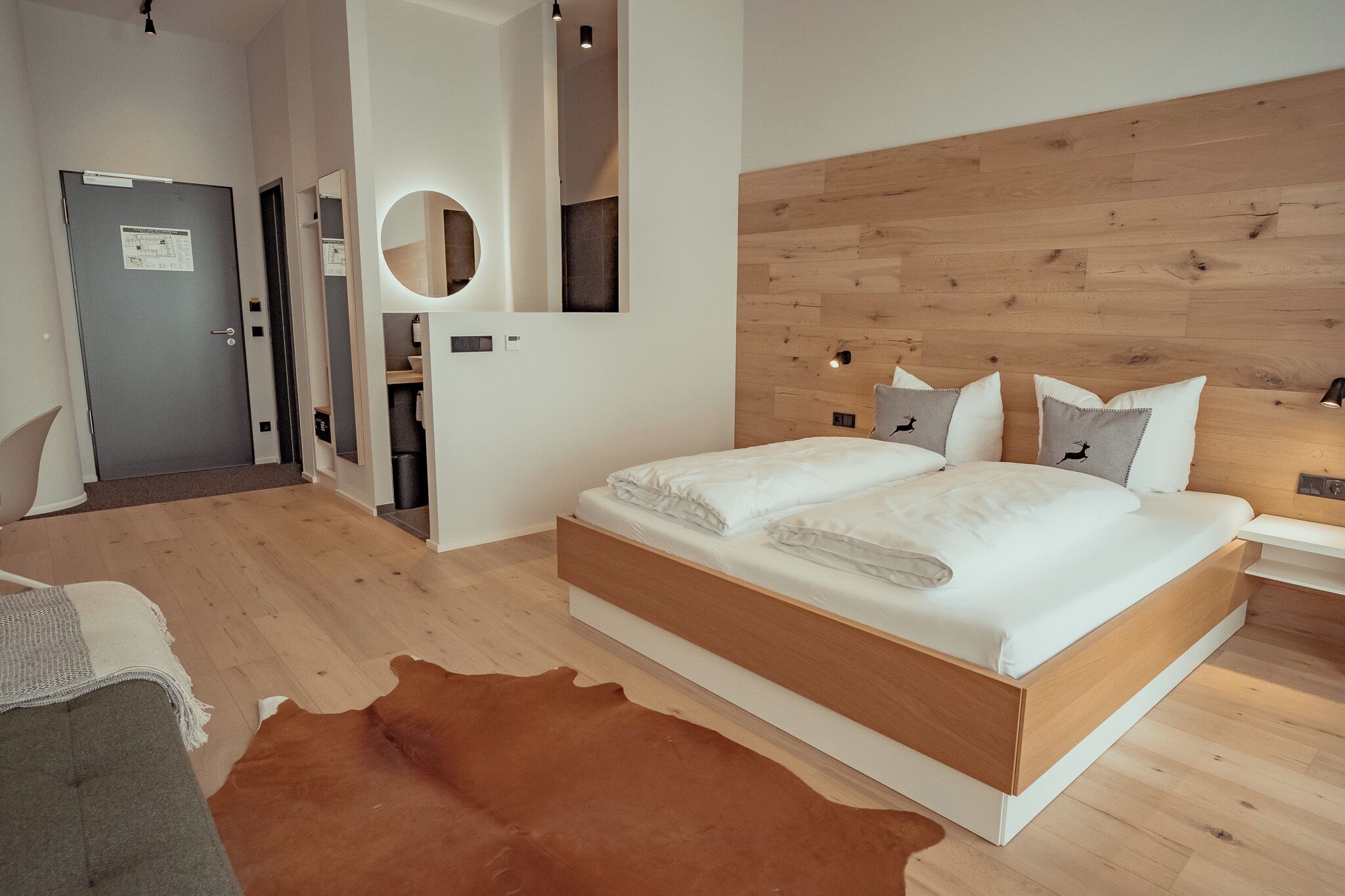 Un dormitorio moderno con cabecera de madera, una cama grande con ropa blanca, un espejo redondo y un tapete de cuero de vaca.