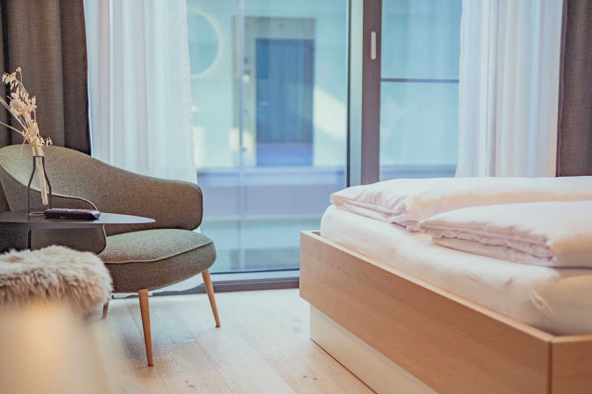 A modern hotel room with a wooden bed, a grey armchair, a small table with a vase, and a large window offering a view of the outside.