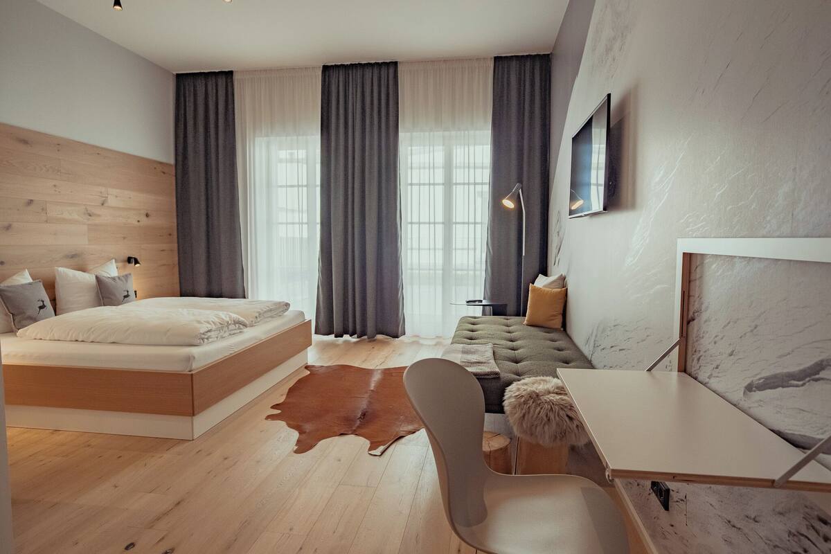 A modern bedroom with a wooden floor, a large bed, a sofa, a desk, and a leather rug.